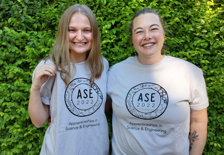 ASE 2022 t-shirt unveiling featuring ASE Program Manager Maura and ASE Program Coordinator Allison sporting this year's design on an icy grey tee. The design is printed in black and is a circle with various STEM-related icons shown inside with "ASE 2022"