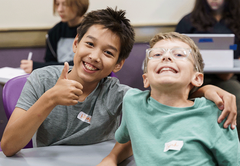 Two kids posing together for a photo, one is holding a thumb up and smiling while the other smiles.