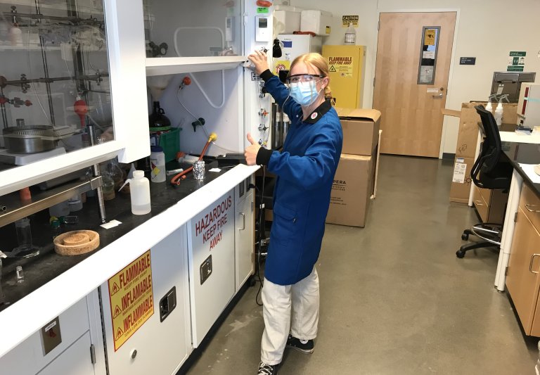 young adult scientist working in a chemistry lab at the fume hood, wearing a lab coat and safety glasses and giving a thumbs up