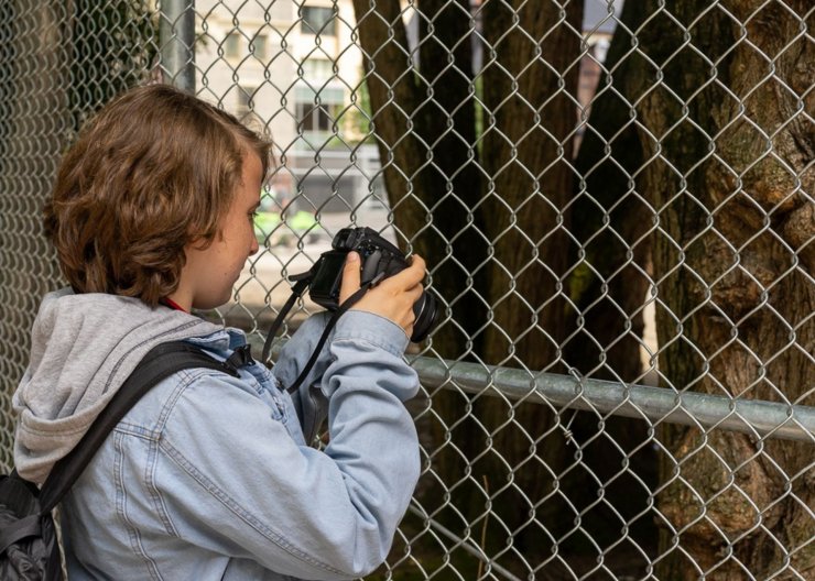 A young Saturday Academy student stands in front of a chainlink fence checking out their DSLR camera in our Digital Photography summer camp 