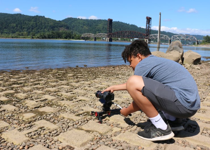 A student, outside on a sunny day, lining up a video camera on the Willamette River in Saturday Academy’s Digital Video Making camp