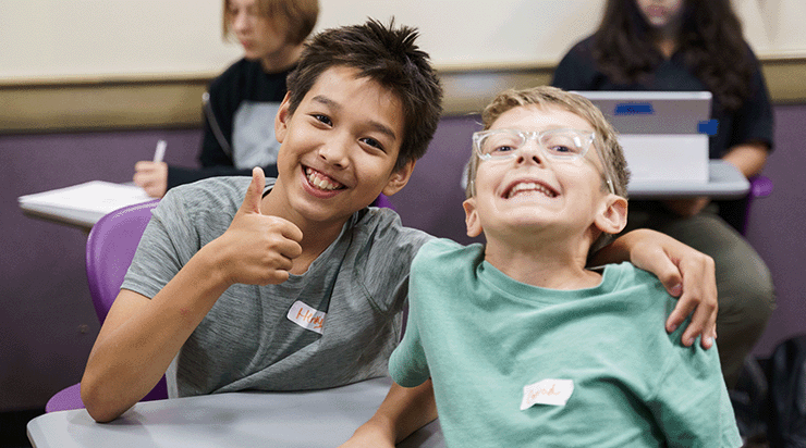Two kids posing together for a photo, one is holding a thumb up and smiling while the other smiles.