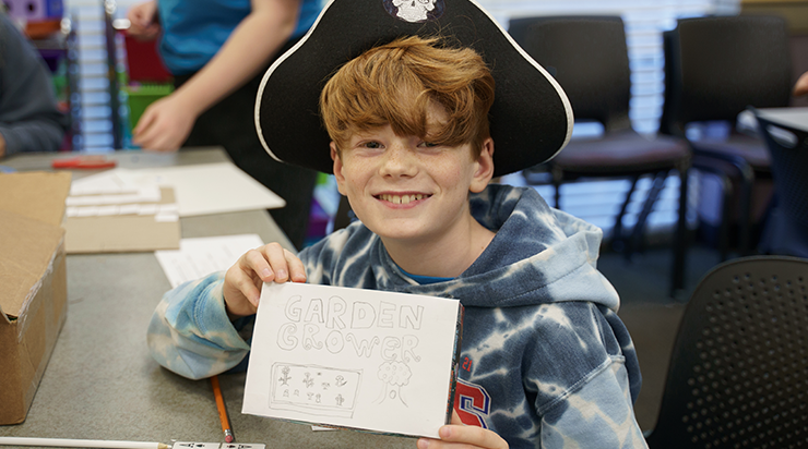 Board Game student showing off their board game box design that says 'Garden Grower'.